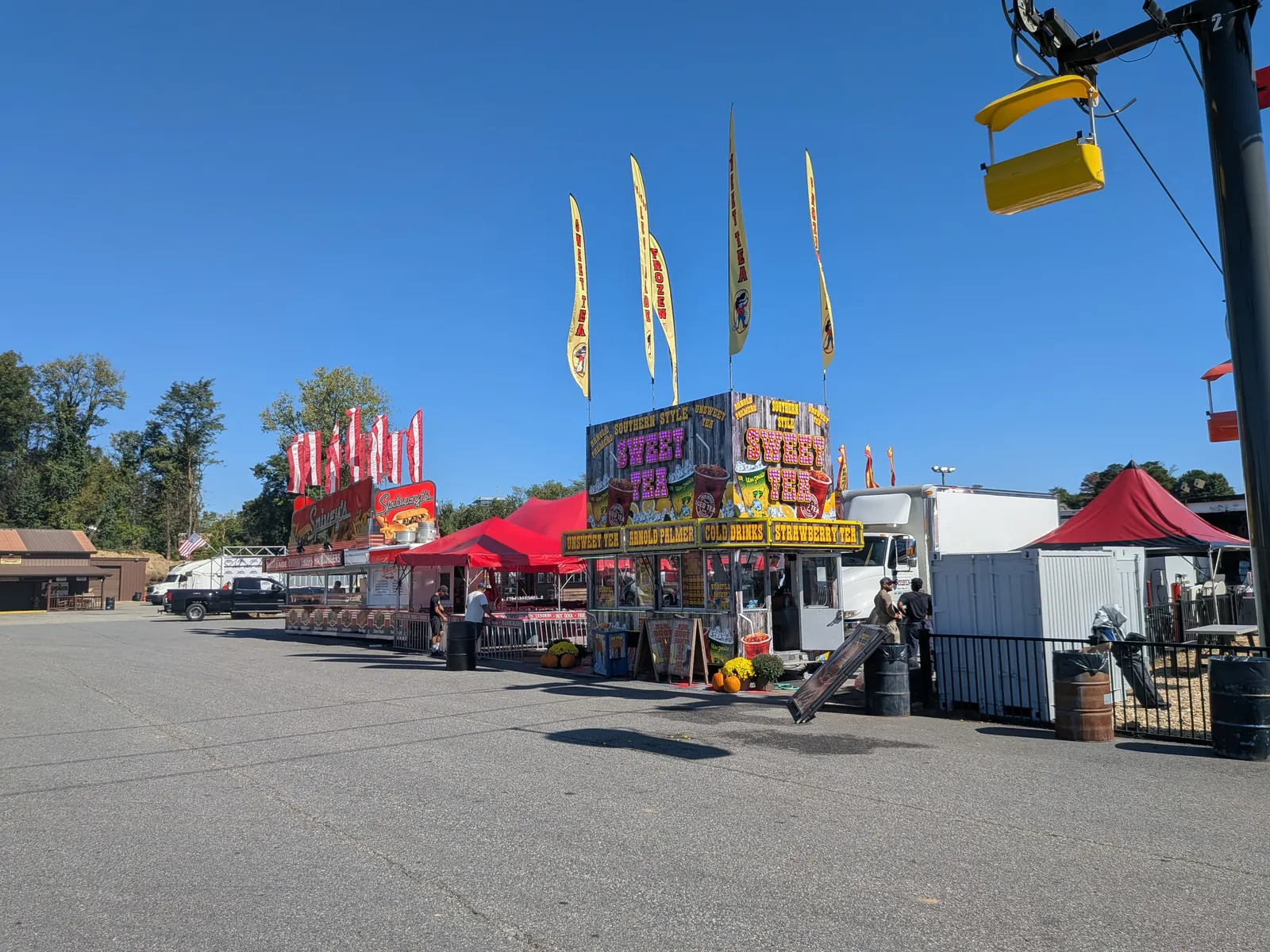 Food stands with banner flags, including a sweet tea stand, on an asphalt lot, with a sky-ride chair overhead at right.