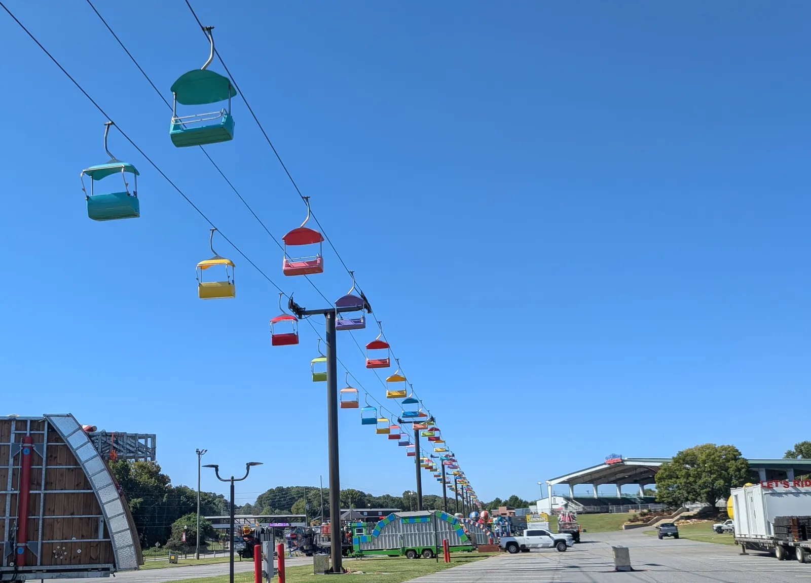 Rows of colorful sky-ride chairs hanging from cables over the fairgrounds, with a covered grandstand and parked trailers in the distance.