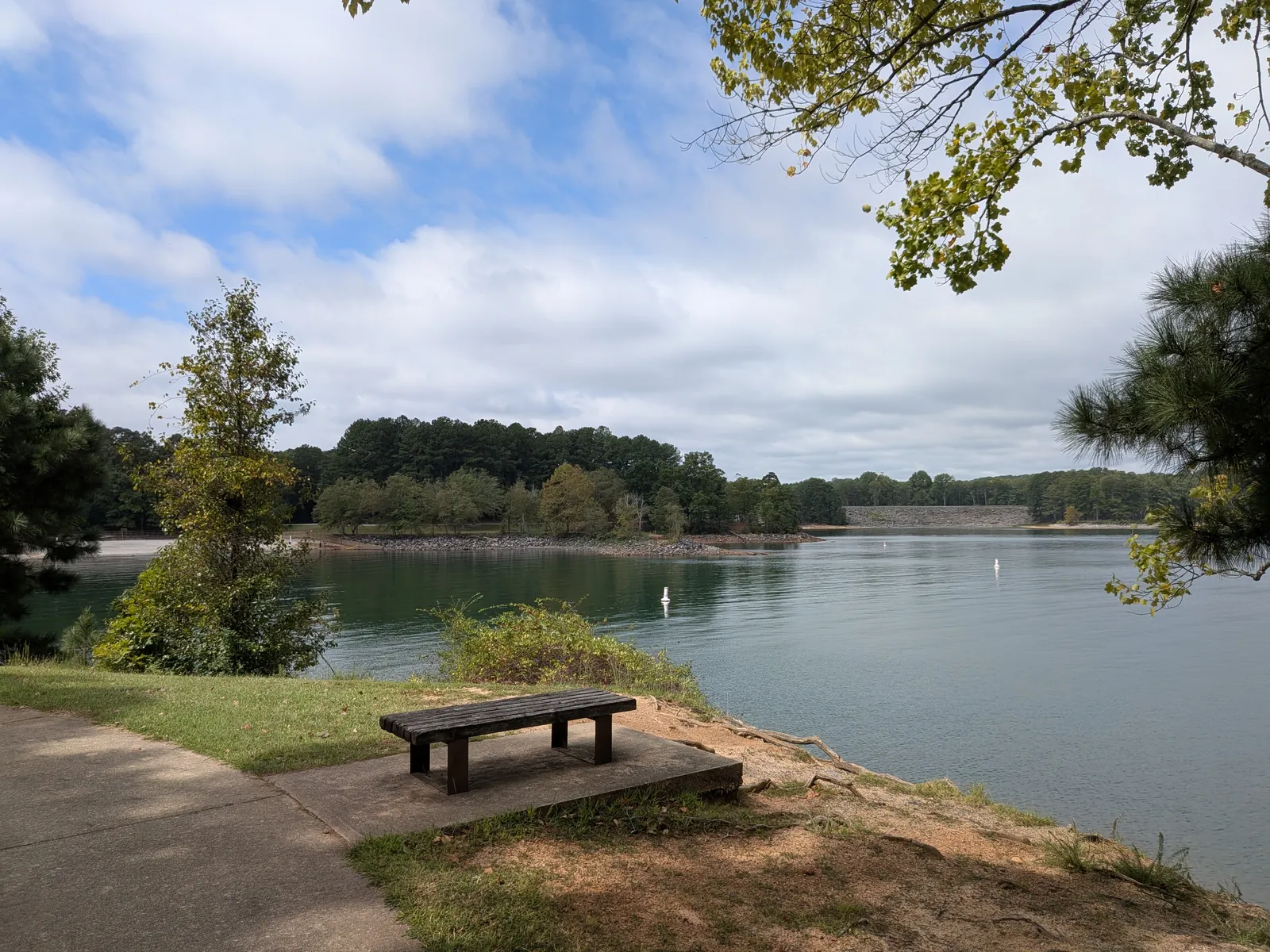 Lake Lanier seen from a bench beside a paved path at West Bank Park, with a wooded shoreline across calm water under a partly cloudy sky